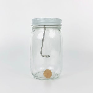 May include: A clear glass jar with a white lid and a metal hook inside. A small, round, wooden cork sits at the bottom of the jar.