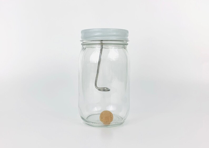 May include: A clear glass jar with a white lid and a metal hook inside. A small, round, wooden cork sits at the bottom of the jar.