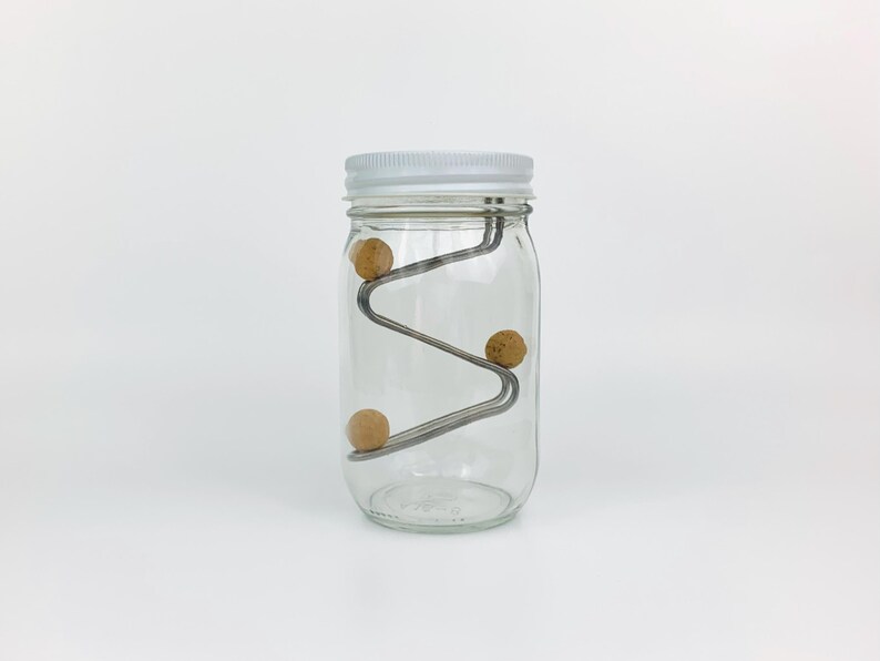 May include: A clear glass jar with a white lid contains a metal wire structure with three wooden balls rolling down the structure.