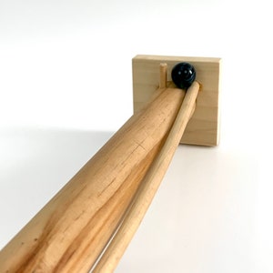 May include: A wooden toy with a marble rolling down a track. The track is made of two wooden dowels and a wooden base. The marble is black.
