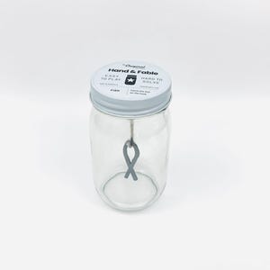 May include: A glass jar with a white lid that says "Hand & Fable" on it. The lid also says "Easy to Play" and "Hard to Solve". There is a gray ribbon inside the jar.
