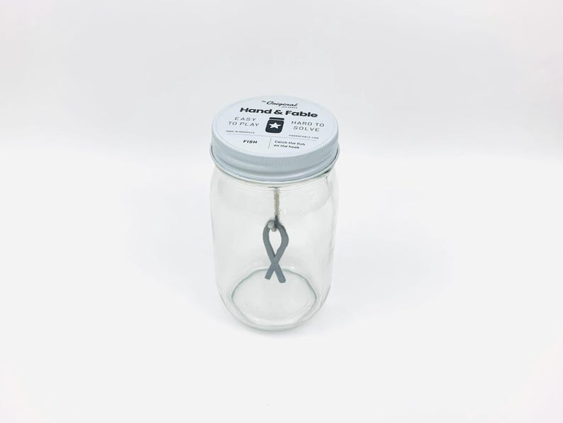 May include: A glass jar with a white lid that says "Hand & Fable" on it. The lid also says "Easy to Play" and "Hard to Solve". There is a gray ribbon inside the jar.