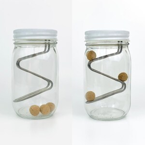 May include: Two glass jars with white lids and a metal wire inside. The wire is shaped like a zig-zag and has small brown balls rolling down it. The jar on the left has three balls at the bottom, while the jar on the right has one ball rolling down the wire.