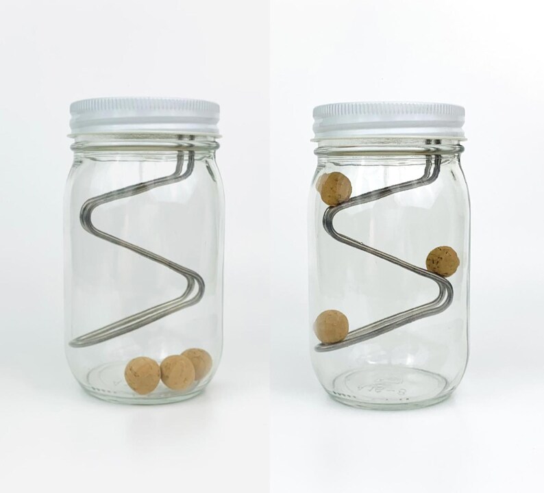 May include: Two glass jars with white lids and a metal wire inside. The wire is shaped like a zig-zag and has small brown balls rolling down it. The jar on the left has three balls at the bottom, while the jar on the right has one ball rolling down the wire.