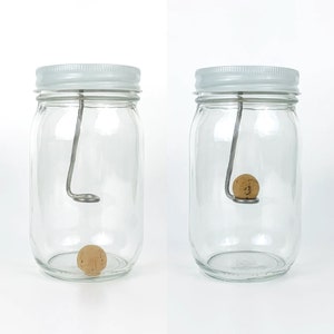 May include: Two clear glass jars with white lids, each containing a cork ball suspended by a metal wire. The wire is bent into a hook shape at the top and is attached to the lid.