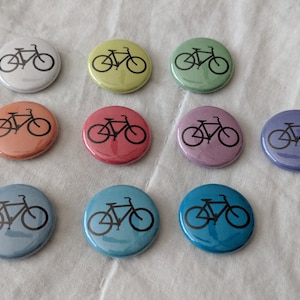 Puede incluir: Una colecci&oacute;n de botones redondos en varios colores, cada uno con un gr&aacute;fico de bicicleta negra. Los botones son en tonos de blanco, amarillo, verde, naranja, rojo, morado y azul. El dise&ntilde;o de la bicicleta es un contorno simple.