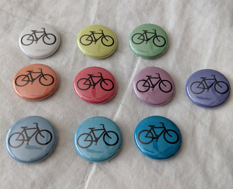 Puede incluir: Una colecci&oacute;n de botones redondos en varios colores, cada uno con un gr&aacute;fico de bicicleta negra. Los botones son en tonos de blanco, amarillo, verde, naranja, rojo, morado y azul. El dise&ntilde;o de la bicicleta es un contorno simple.