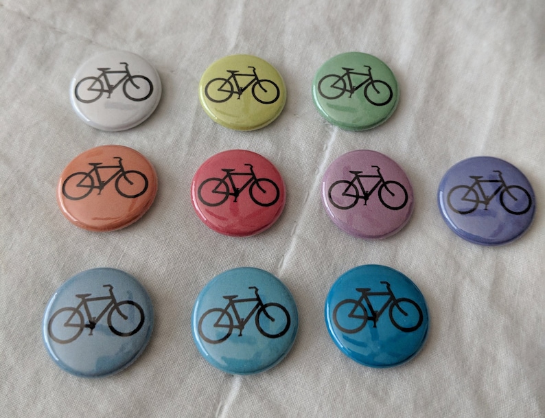 Puede incluir: Un conjunto de diez botones redondos de varios colores, cada uno con un dise&ntilde;o de bicicleta negra. Los botones son de color blanco, amarillo, verde, naranja, rojo, rosa, morado, azul claro y turquesa. Est&aacute;n colocados sobre una tela blanca.