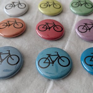 Puede incluir: Una colecci&oacute;n de botones redondos y coloridos, cada uno con una silueta de bicicleta negra. Los botones son de varios tonos pastel, incluyendo rosa, azul, verde y amarillo. Est&aacute;n dispuestos sobre una tela blanca.