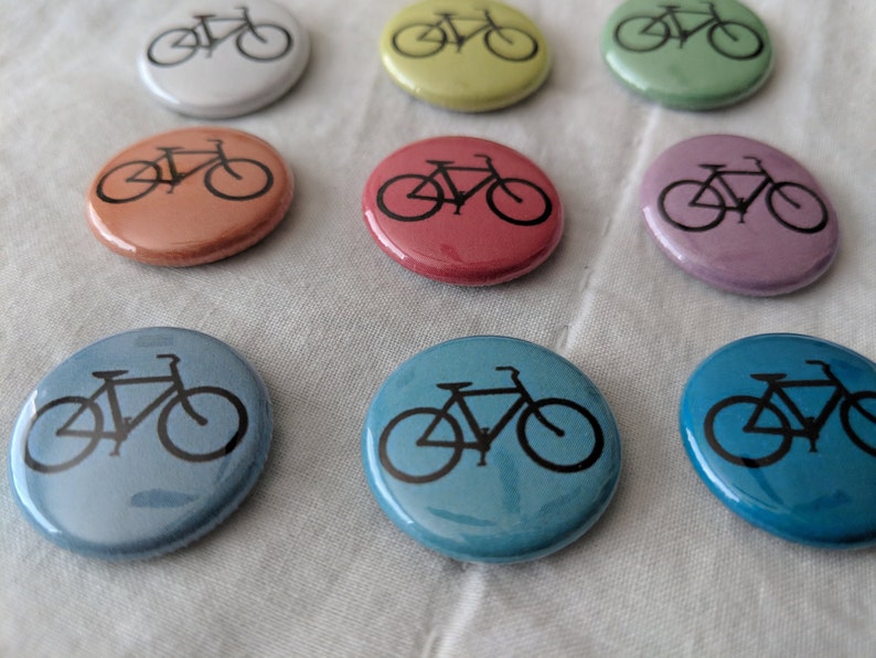 Puede incluir: Una colecci&oacute;n de botones redondos y coloridos, cada uno con una silueta de bicicleta negra. Los botones son de varios tonos pastel, incluyendo rosa, azul, verde y amarillo. Est&aacute;n dispuestos sobre una tela blanca.