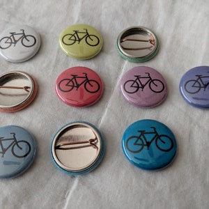 Puede incluir: Un conjunto de botones redondos con diferentes colores, cada uno con una silueta negra de bicicleta. Los colores incluyen blanco, amarillo-verdoso, rojo, morado, azul y turquesa. Los botones miden aproximadamente 2,5 cm de di&aacute;metro.