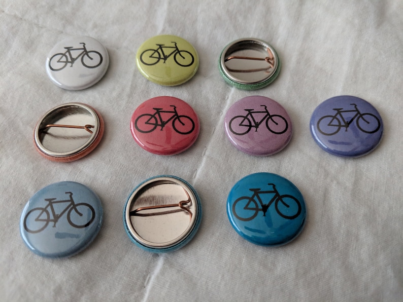 Puede incluir: Un conjunto de botones redondos con diferentes colores, cada uno con una silueta negra de bicicleta. Los colores incluyen blanco, amarillo-verdoso, rojo, morado, azul y turquesa. Los botones miden aproximadamente 2,5 cm de di&aacute;metro.