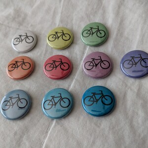 Puede incluir: Una colecci&oacute;n de diez botones redondos en varios colores, cada uno con una silueta de bicicleta negra. Los botones var&iacute;an en color desde el blanco hasta el azul, con tonos de amarillo, naranja, rojo y morado. Los botones est&aacute;n dispuestos sobre una superficie blanca texturizada.