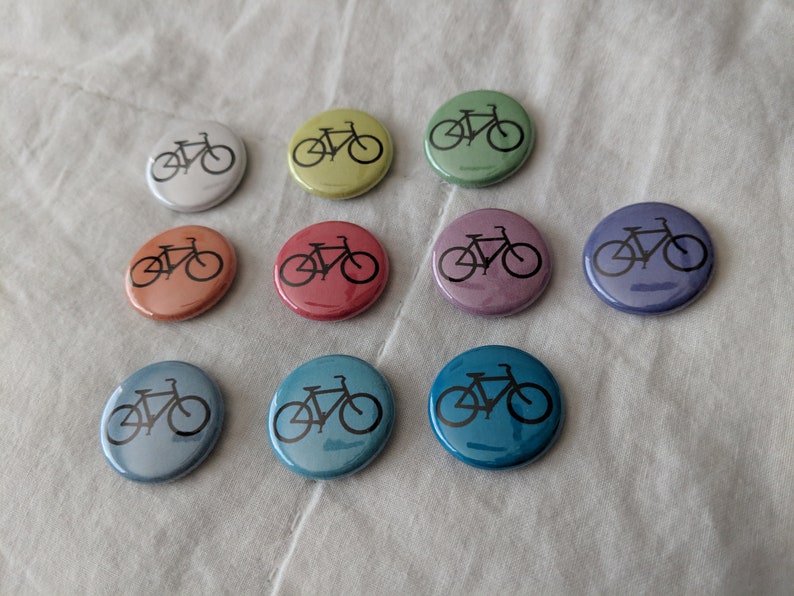 Puede incluir: Una colecci&oacute;n de diez botones redondos en varios colores, cada uno con una silueta de bicicleta negra. Los botones var&iacute;an en color desde el blanco hasta el azul, con tonos de amarillo, naranja, rojo y morado. Los botones est&aacute;n dispuestos sobre una superficie blanca texturizada.