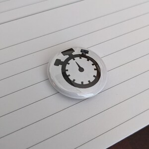 May include: A white button with a black stopwatch design. The stopwatch has a circular face with a needle pointing to the time. The button is round and glossy.