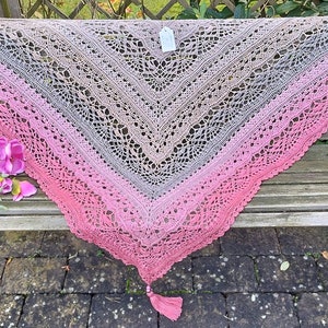 May include: A crocheted shawl with a pink, gray, and brown ombre pattern. The shawl is draped over a wooden bench with a floral accent. The shawl has a tassel at the bottom.