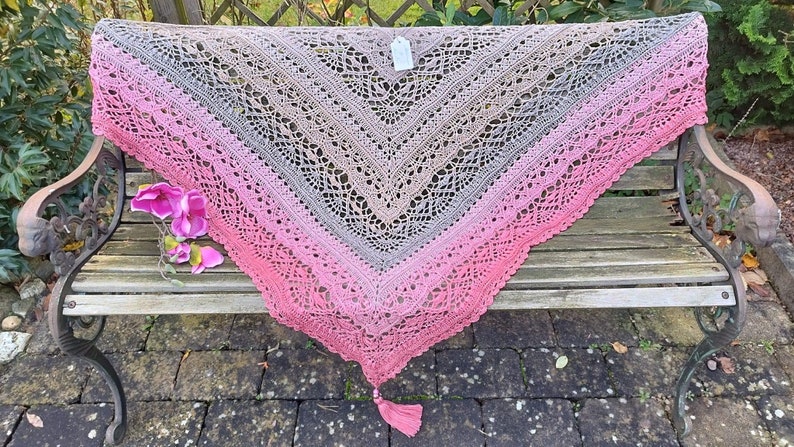 May include: A crocheted shawl with a pink, gray, and brown ombre pattern. The shawl is draped over a wooden bench with a floral accent. The shawl has a tassel at the bottom.