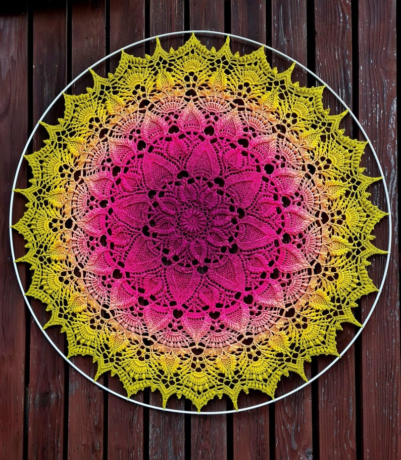Puede incluir: Un gran doily circular de ganchillo con un dise&ntilde;o degradado de rosa y amarillo. El doily est&aacute; hecho con puntadas delicadas e intrincadas y tiene un borde festoneado.