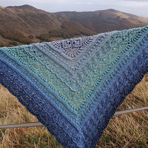 May include: A blue and green crocheted shawl with a lacy pattern, draped over a fence against a backdrop of mountains.