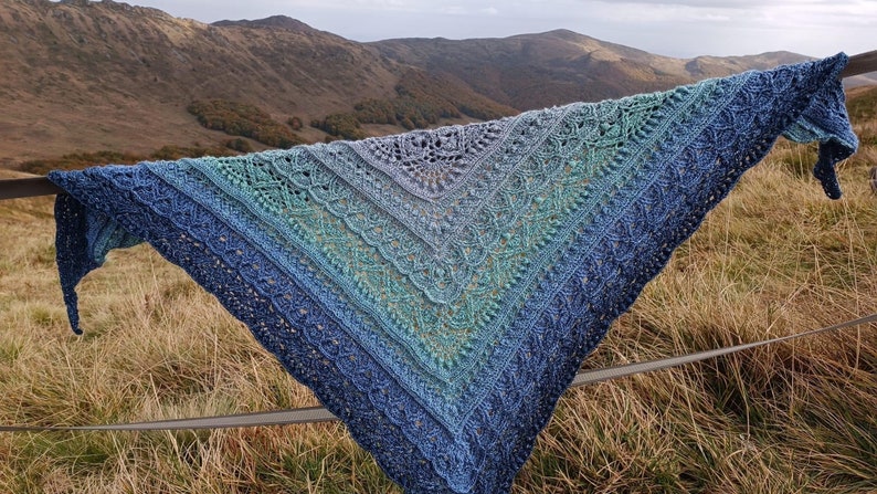 May include: A blue and green crocheted shawl with a lacy pattern, draped over a fence against a backdrop of mountains.