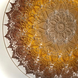 Puede incluir: Un gran doily redondo de ganchillo con un degradado de color marr&oacute;n a dorado. El doily est&aacute; colgado en una pared blanca.