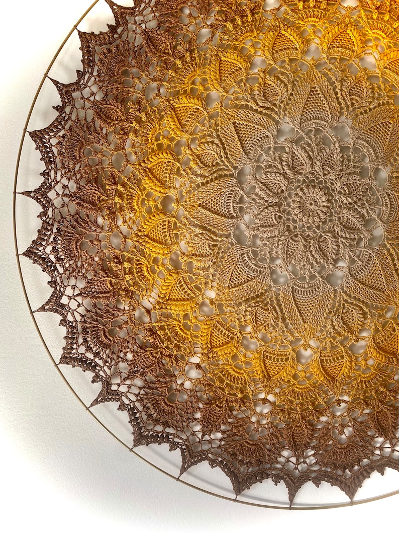 Puede incluir: Un gran doily redondo de ganchillo con un degradado de color marr&oacute;n a dorado. El doily est&aacute; colgado en una pared blanca.