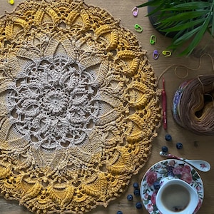 Puede incluir: Un mandala tejido a crochet con un degradado de colores amarillo y marr&oacute;n. El mandala est&aacute; colocado sobre una superficie de madera con una taza de caf&eacute; y ar&aacute;ndanos.