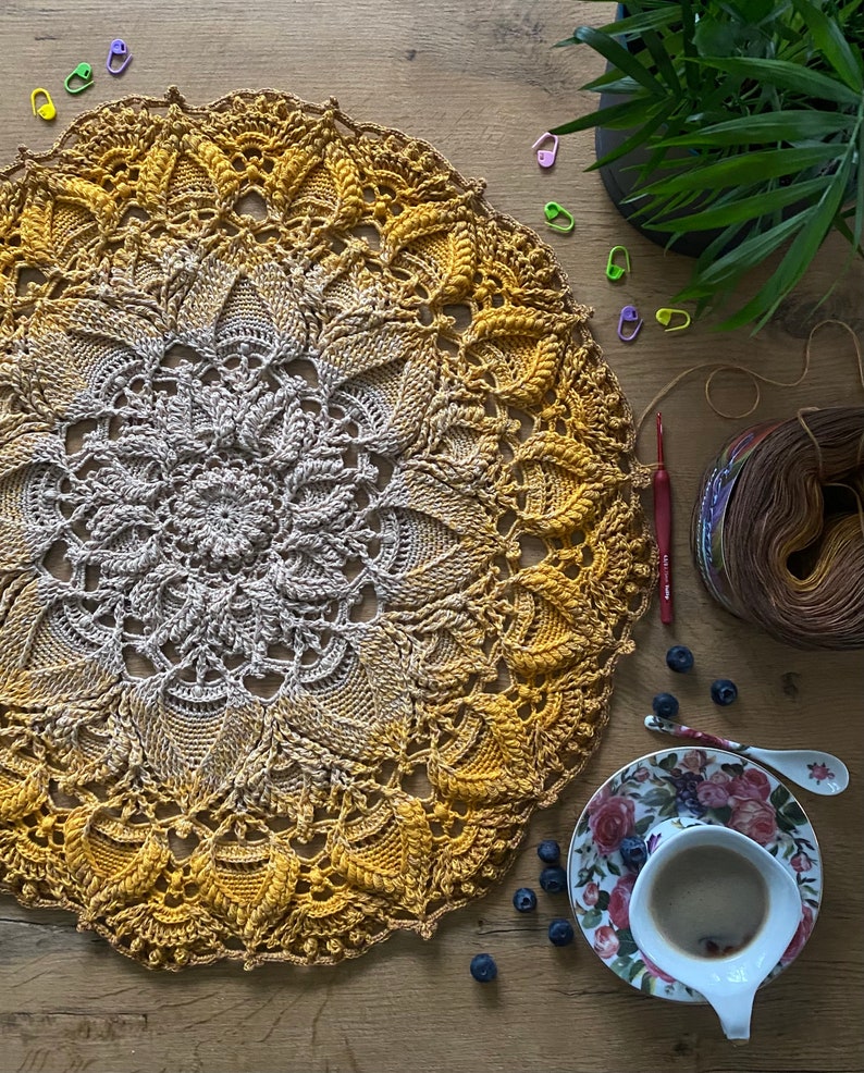 Puede incluir: Un mandala tejido a crochet con un degradado de colores amarillo y marr&oacute;n. El mandala est&aacute; colocado sobre una superficie de madera con una taza de caf&eacute; y ar&aacute;ndanos.