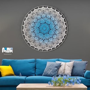 Puede incluir: Un gran tapiz de pared de macram&eacute; blanco con un efecto degradado azul. El mandala est&aacute; colgado en una pared gris sobre un sof&aacute; azul con almohadas. El sof&aacute; est&aacute; en un ambiente de sala de estar con una mesa de caf&eacute; y plantas.
