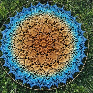 Puede incluir: Un gran mandala tejido a crochet con un dise&ntilde;o degradado en azul y marr&oacute;n. El mandala es circular y tiene un patr&oacute;n intrincado de flores y hojas. Se muestra sobre un fondo de hierba verde.