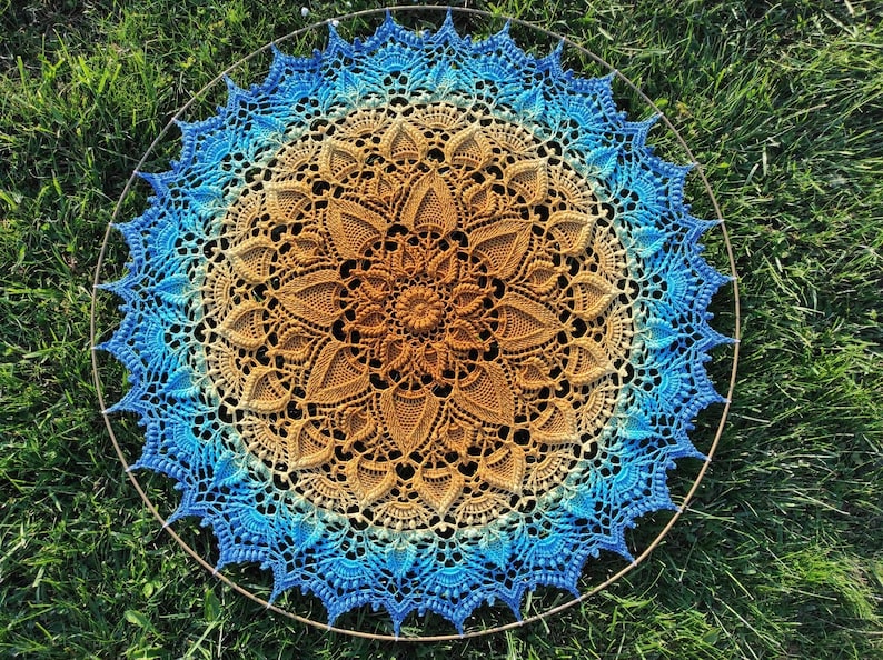 Puede incluir: Un gran mandala tejido a crochet con un dise&ntilde;o degradado en azul y marr&oacute;n. El mandala es circular y tiene un patr&oacute;n intrincado de flores y hojas. Se muestra sobre un fondo de hierba verde.