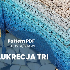 Pode incluir: Um xale de crochê com um degradê de cores azul e branco. O xale está drapeado sobre uma pilha de toras de madeira. O texto "Pattern PDF CHUSTA/SHAWL LUKRECJA TRI" está impresso na imagem.