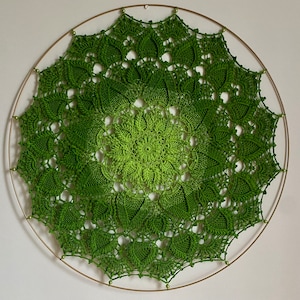 Puede incluir: Un mandala de ganchillo verde con un dise&ntilde;o circular, colgado de un aro de metal dorado.