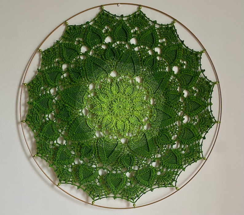 Puede incluir: Un mandala de ganchillo verde con un dise&ntilde;o circular, colgado de un aro de metal dorado.