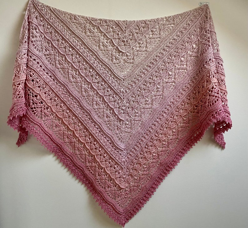 May include: A delicate crocheted shawl with a gradient of pink and beige colors. The shawl has a lacy pattern and a scalloped edge.