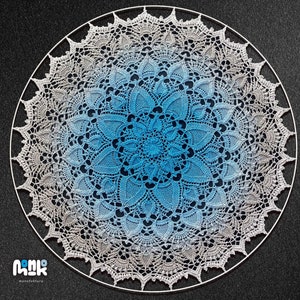 Puede incluir: Un gran doily circular de ganchillo con un degradado de colores azul y blanco. El doily est&aacute; hecho con puntadas delicadas y tiene un dise&ntilde;o intrincado. Se muestra sobre un fondo gris oscuro.