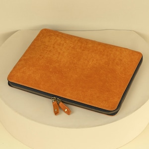 Könnte beinhalten: Eine braune Ledertasche für Laptops mit Reißverschluss. Die Tasche hat eine strukturierte Oberfläche und einen dunkelblauen Reißverschluss.