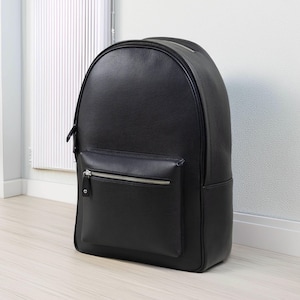 May include: A black leather backpack with a rounded top and a front pocket. The backpack has a zip closure on the main compartment and the front pocket. The bag is made of a textured leather material.