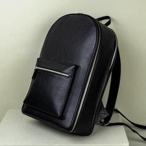 Peut inclure: Un sac à dos noir avec un dessus arrondi et une poche avant. Le sac à dos a une fermeture éclair argentée qui longe le côté et autour de la poche avant. Les bretelles sont noires et fixées au bas du sac.