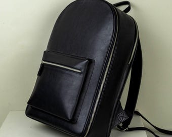 Könnte beinhalten: Ein schwarzer Rucksack mit abgerundeter Oberseite und einer Vordertasche. Der Rucksack hat einen silbernen Reißverschluss, der entlang der Seite und um die Vordertasche verläuft. Die Träger sind schwarz und am Boden der Tasche befestigt.