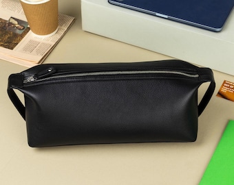 May include: A black leather bum bag with a zip closure, on a neutral surface. A coffee cup, newspaper, tablet, pen, and green notebook are also visible. The bag is approximately 30 cm in length.