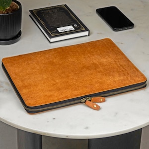 May include: A rectangular laptop sleeve in a textured tan brown finish with a black zip closure. The sleeve features two matching tan brown leather zip pulls with brass-toned metal hardware, providing a secure and functional design.