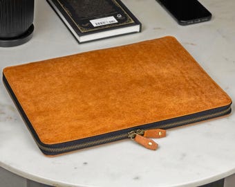 May include: A rectangular laptop sleeve in a textured tan brown finish with a black zip closure. The sleeve features two matching tan brown leather zip pulls with brass-toned metal hardware, providing a secure and functional design.