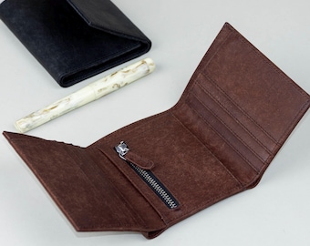 Könnte beinhalten: Offene braune Lederbrieftasche mit Reißverschlusstasche und mehreren Kartenfächern. Eine schwarze Brieftasche und ein Stift mit weißem und beigem Muster sind ebenfalls zu sehen. Die Brieftasche ist geöffnet und zeigt ihr Inneres.