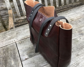 Puede incluir: Un bolso tote de cuero burdeos con correas de cuero negro y herrajes plateados. El bolso está sobre una superficie de madera.