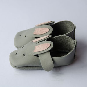 May include: A pair of gray leather baby shoes with pink felt bunny ears. The shoes have a soft, flexible sole and are perfect for crawling and walking.