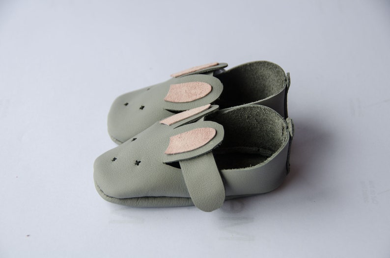 May include: A pair of gray leather baby shoes with pink felt bunny ears. The shoes have a soft, flexible sole and are perfect for crawling and walking.