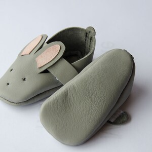 May include: A pair of soft, gray leather baby shoes with bunny ears. The shoes have a strap across the top and are made of a soft, flexible material.