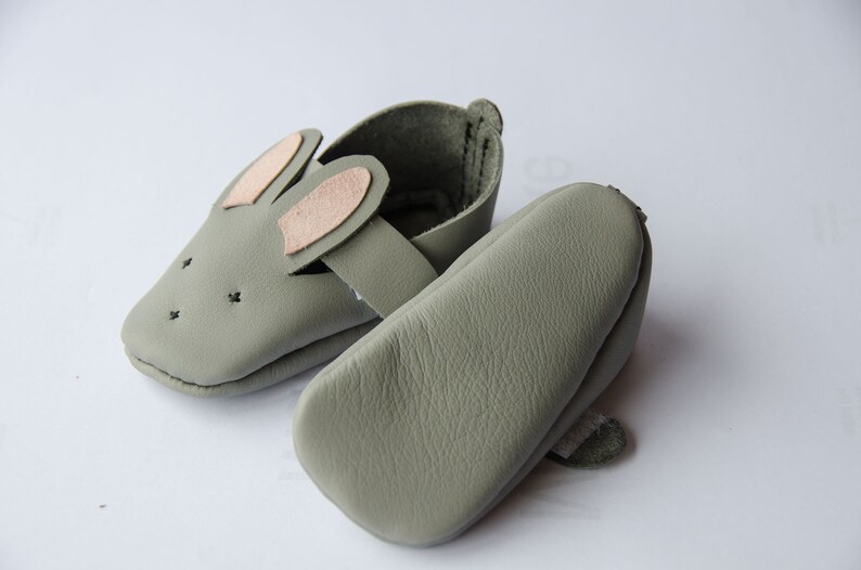 May include: A pair of soft, gray leather baby shoes with bunny ears. The shoes have a strap across the top and are made of a soft, flexible material.