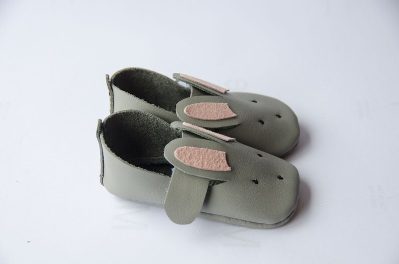 May include: A pair of green leather baby shoes with pink bunny ears. The shoes have a soft, flexible sole and are perfect for little feet.